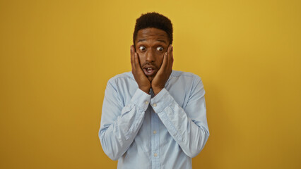 Obraz premium Young man with hands on face stands surprised against an isolated yellow background wearing a light blue shirt showcasing expressive emotions and appealing features
