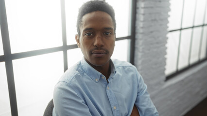 Fototapeta premium Young man in office setting with natural light through large windows exudes confidence and professionalism, seated casually in a modern workspace with a contemplative expression.