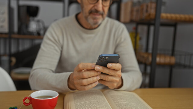 Mature man using smartphone while sitting at a desk with a red coffee cup in an office environment, with an open book, showcasing a calm workplace setting.