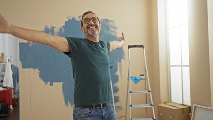 Mature hispanic man smiling with arms outstretched in a newly painted living room, with a ladder and paint supplies, enjoying the fresh feeling of his new home. © Krakenimages.com