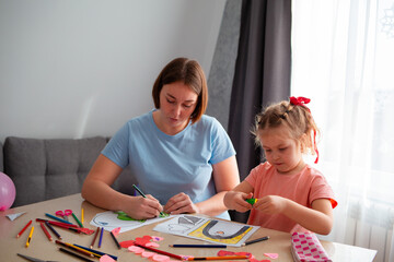 Obraz premium Wide shot of Caucasian woman teacher and little cute girl decorate Valentine's Day cards together, sitting at the table. Mother and daughter's joint leisure time