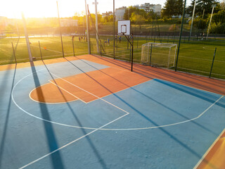 Outdoor basketball court at sunset with long shadows and warm light, adjacent to a soccer field where players are training, creating a dynamic urban sports atmosphere © sandsun