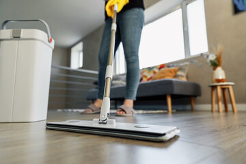 Woman mopping, wiping sleek hardwood floor in modern home setting. Girl wearing yellow gloves. Housewife uses professional cleaning supplies: mop, plastic bucket with wringer in cozy living house room
