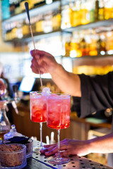 A bartender stirs two red cocktails in tall glasses with ice at a bar. The background features...