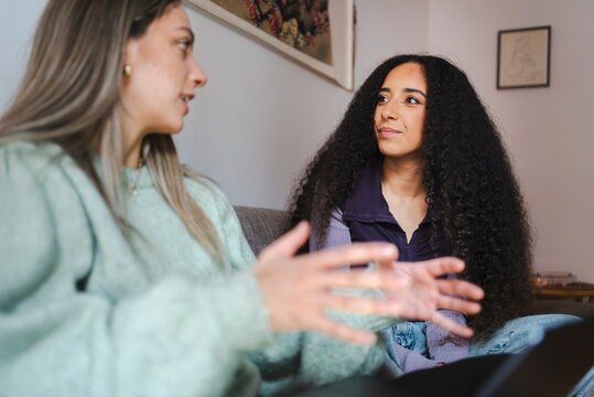 A woman in a mint green sweater gestures while speaking to her curly-haired friend, who listens intently, as they share a meaningful conversation on a cozy couch at home.