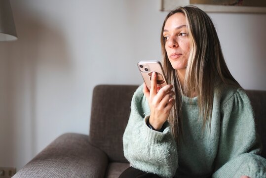 A young woman in a mint green sweater sits on a couch, holding her phone and gazing thoughtfully into the distance, appearing deep in thought in a cozy home setting.