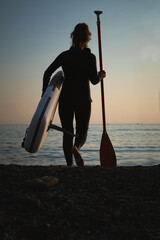 Old aged woman walking with surf board and oar, sunset on sea. Active lifestyle for older senior people. Elderly female make sport at ocean, surfing. Summer holidays vacation travel. SUP paddle
