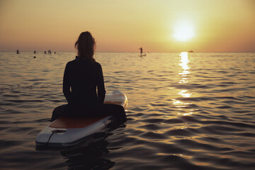 An elderly woman rides paddleboarding in the ocean. Senior female is waiting for a wave on a surfboard. Active old age. Old girl is sitting on a board in the sea at sunset, dressed in a swimming suit