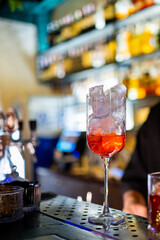 A close-up of a cocktail glass filled with ice and a red beverage, placed on a bar counter with blurred bottles in the background, creating a vibrant and inviting atmosphere.
