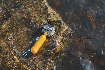 An action camera with a waterproof case and a float for swimming rests on a rock in the water.