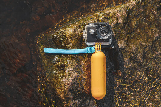 The action camera lies on a stone with a yellow float and a waterproof case in the water. Hobby photo video filming.