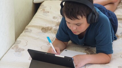 Portrait of a boy using tablet computer and headphone during pandemic lockdown, homeschooling,...