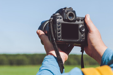 Close-up of a professional camera in the hands of a man. Background of green field and blue sky.