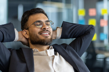Successful businessman resting while sitting at his desk inside the office. Well done man with his hands behind his head with closed eyes smiling, satisfied with results of achieving the workplace.
