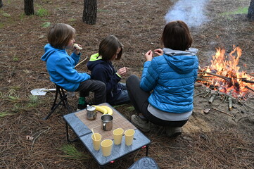 Mother and two children sit by a campfire in the forest, eating and relaxing at a small table. They enjoy a cozy outdoor meal and family time surrounded by pine trees and nature