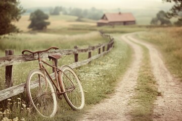 Rustic rusty bicycle leans against a wooden fence beside a winding country road leading to a farmhouse in a misty field.