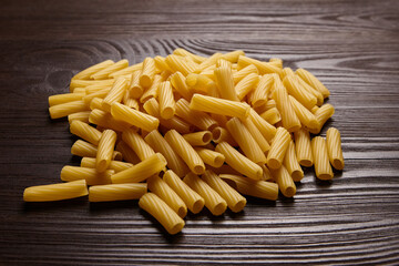 Uncooked tortiglioni pasta spread on a dark wood table, featuring cylindrical ridged shapes and golden-yellow texture under soft natural lighting