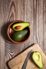 One halved avocado with seed and one whole in a wooden bowl, another half and knife on a cutting...