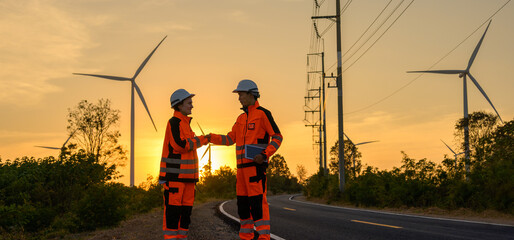 Two engineers in bright orange safety uniform and white helmet shaking hands beside road in wind farm at sunset with golden sky. Teamwork successful and renewable green energy development concept