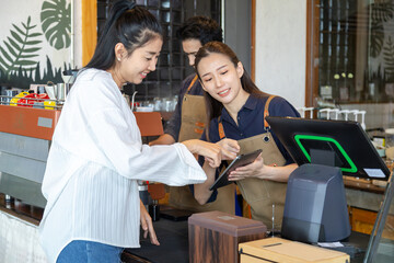 Asian cafe cashier worker female showing menu or cashless billing payment to customer, barista small business owner using technology tablet receives coffee order at workplace in restaurant coffee shop