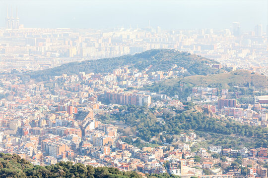 Panoramic view of Barcelona blending urban density with green hill under a hazy sky. City stretches toward the Balearic coast, showcasing its vibrant mix of nature and modern architecture