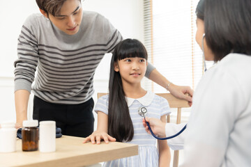 Asian female doctor smiling while examining a young girl, her father standing nearby .Healthcare worker checking the health of a patient girl.