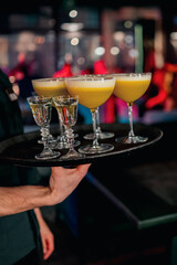 A waiter holds a tray with three elegant yellow cocktails and three ornate clear shots, set against a blurred bar or nightclub background, creating a lively atmosphere.