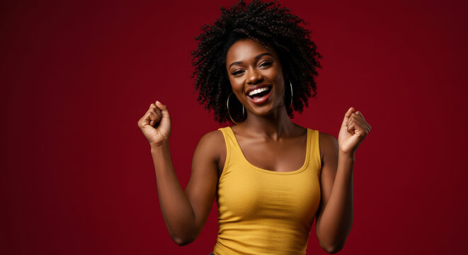 Happy young woman in a mustard yellow tank top celebrating with raised fists against a deep red background. African American female professional in a studio setting.