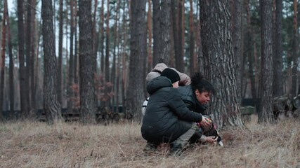 A woman of a multiracial background, accompanied by school-aged boys, is seen crouching and standing in a forest while interacting with a small dog during a brisk winter walk.