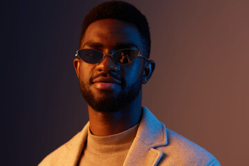 Obraz premium Stylish young man in sunglasses wearing a beige turtleneck and a light gray coat, showing confidence against a gradient background, trendy fashion concept