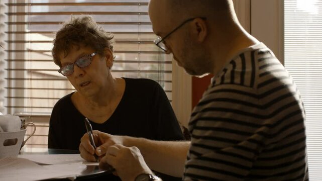 An elderly woman sits at a kitchen table with a younger man. They are reviewing, and signing documents together. The man signs first, followed by the woman. Both wear reading glasses. Warmly sun lit.