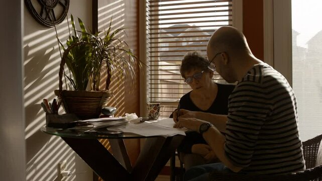 An elderly mother sits at a kitchen table with adult son, in a warmly sunlit house . He helps her fill out a paper questionnaire. She answers, and they discuss. Both wear reading glasses. 24fps.