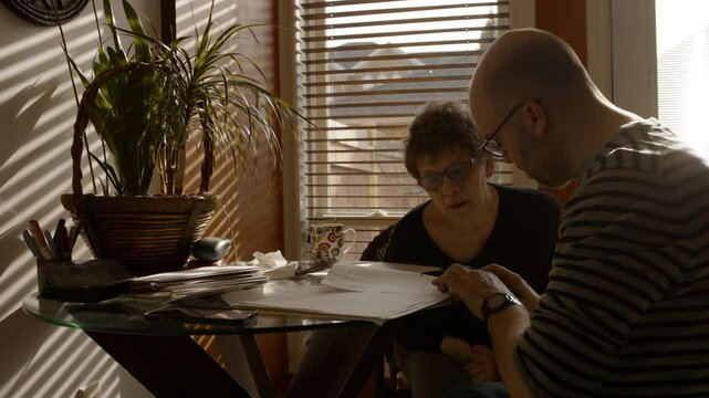 A senior woman sits at a kitchen table with a middle-aged man, in a warmly sunlit house . He helps her fill out a paper questionnaire. She answers, as he writes with pen. Both wear glasses. 24fps.