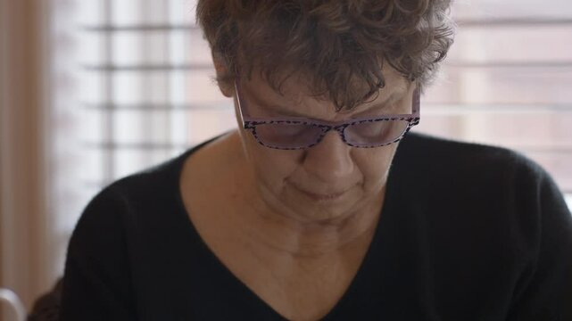 An elderly woman's face focused on gently self-massaging the spasticity in her hemiplegic right hand. Seated by round glass table with legs in checkered pants, Camera tilts down from face to hands.