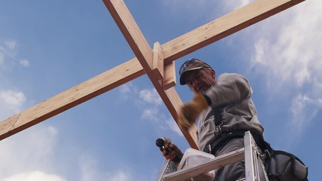 A carpenter working on a construction project stands on a ladder and then screws a small board into an overhead cross support against a bright blue sky.