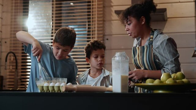 An adult Black woman is teaching her two school-age sons how to cook; one of the boys is whisking ingredients, while the other adds flour to the bowl, surrounded by apples and ingredients.