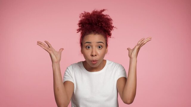 Portrait shocked African American redhead curly woman showing mind explosion gesture, mind blown of ideas looking camera. Female in white t-shirt standing on isolated pink background with copy space