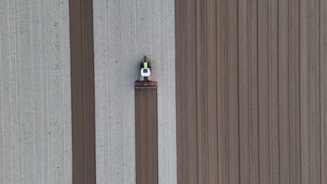 Aerial top down view of a tractor using a rotary tiller to break up compacted soil into evenly spaced rows in northern Italy near the Po River, preparing the field for seeding, real time drone shot