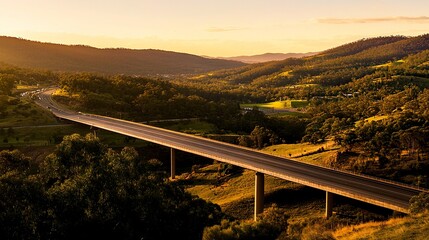 Elevated shot of a motorway bridge at sunset, spanning a valley of green forests and rolling hills, warm hues accentuating its role in development. 