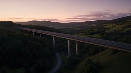 Obraz premium Overhead dusk scene of a motorway bridge connecting towns, spanning a lush valley of hills and forests bathed in gentle twilight tones. 