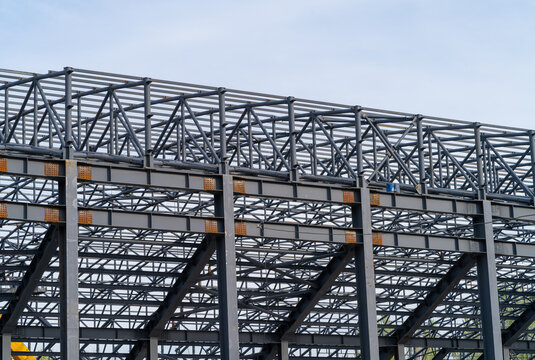 Steel frame grid of exterior walls and arched roof structure of a modular building under construction. Close-up of bolted and welded connections between steel I-beams, supports, and spans.
