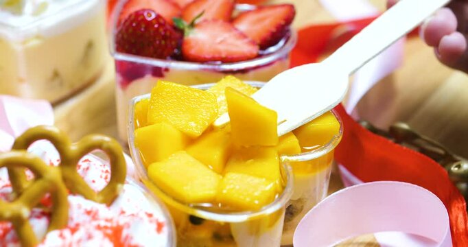 Wooden utensil lifting vibrant mango piece from heart shaped dessert, nestled amid elegant sweets, ribbons, festive decorative elements