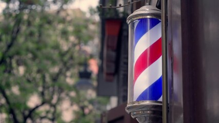 A classic barber pole spins gently in the breeze outside a bustling barbershop, surrounded by urban architecture and greenery.