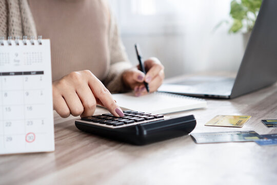Woman calculating monthly debt payments, writing down numbers with a calendar with marked due dates, using a calculator with  credit card on the desk