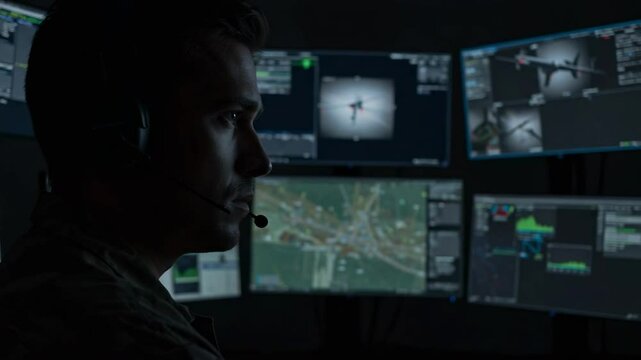 Military operator with headset analyzes drone footage and tactical maps on multiple screens in a dark command center, for surveillance and strategic mission planning operations technology