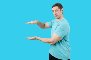 Young man in blue t-shirt holding invisible object with both hands on blue low background. Isolated studio portrait. People and emotions concept. Man pointing at something