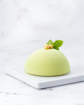Elegant individual green mousse cake with velvet texture, mint leaves and golden pearls. Side view on white marble slab with copy space. Ideal for premium packaging and editorial content.