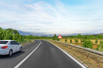 A white sedan drives along a winding countryside road with metal guardrails and a bump warning...