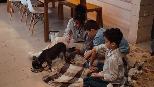 A 30-year-old olive-skinned mother and two school-aged sons sitting on a carpet on the floor and examining a few dog toys and a bottle of water with a drinker next to a French bulldog eating from a bo