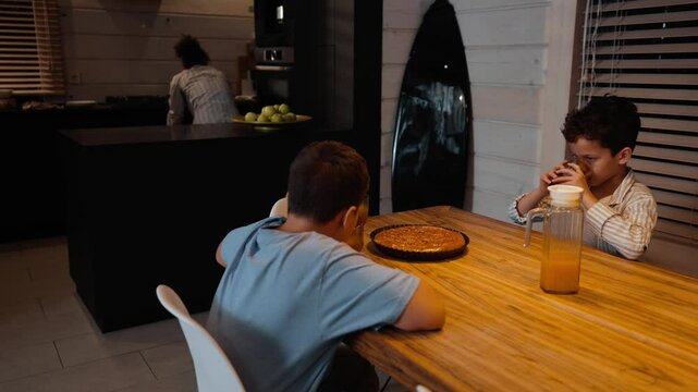 Two brothers sit at the table and drink from glasses while their mother searches through the kitchen drawers